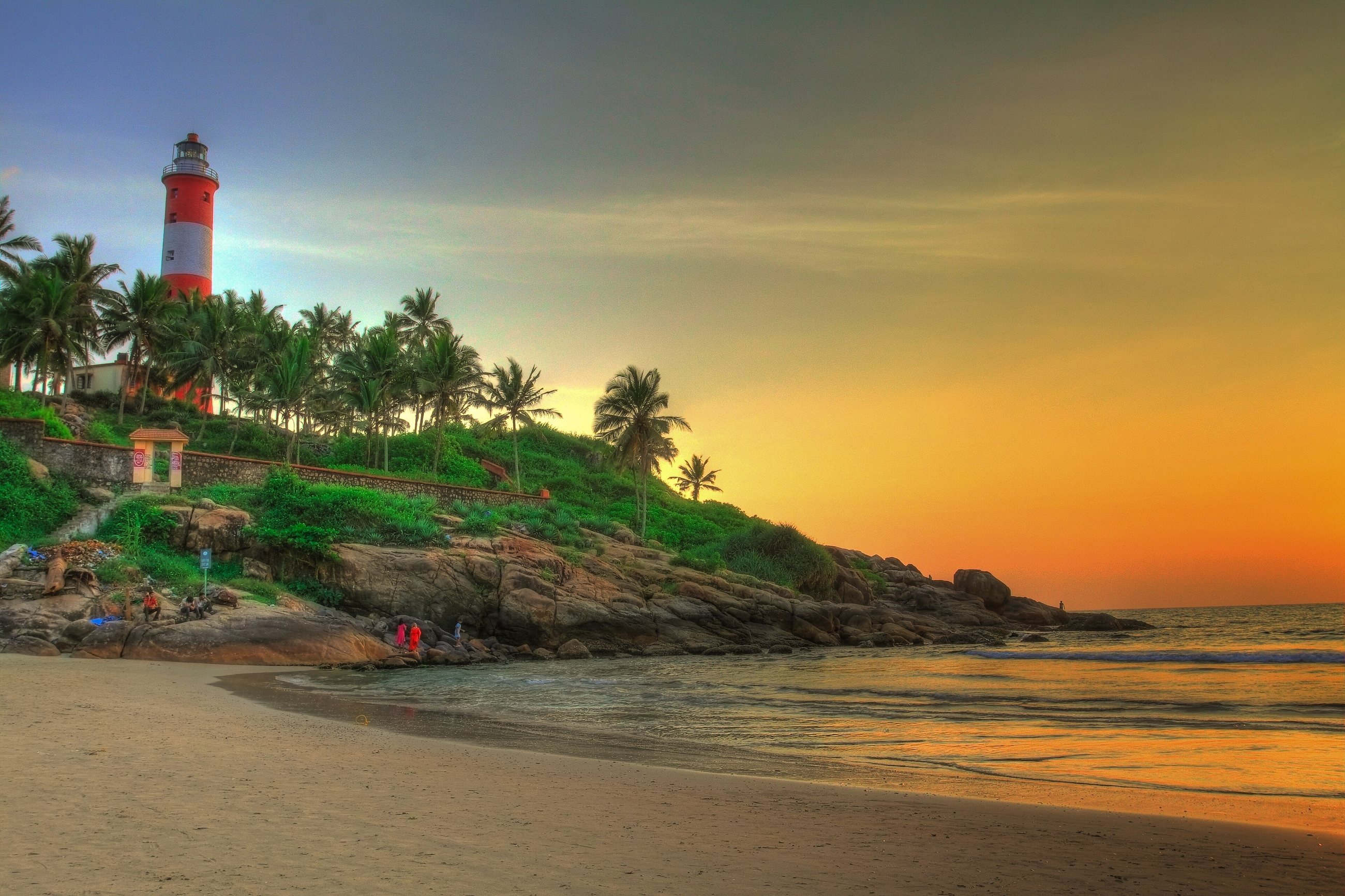 The red and white Kovalam lighthouse above palms and rocks, the beach at sunset