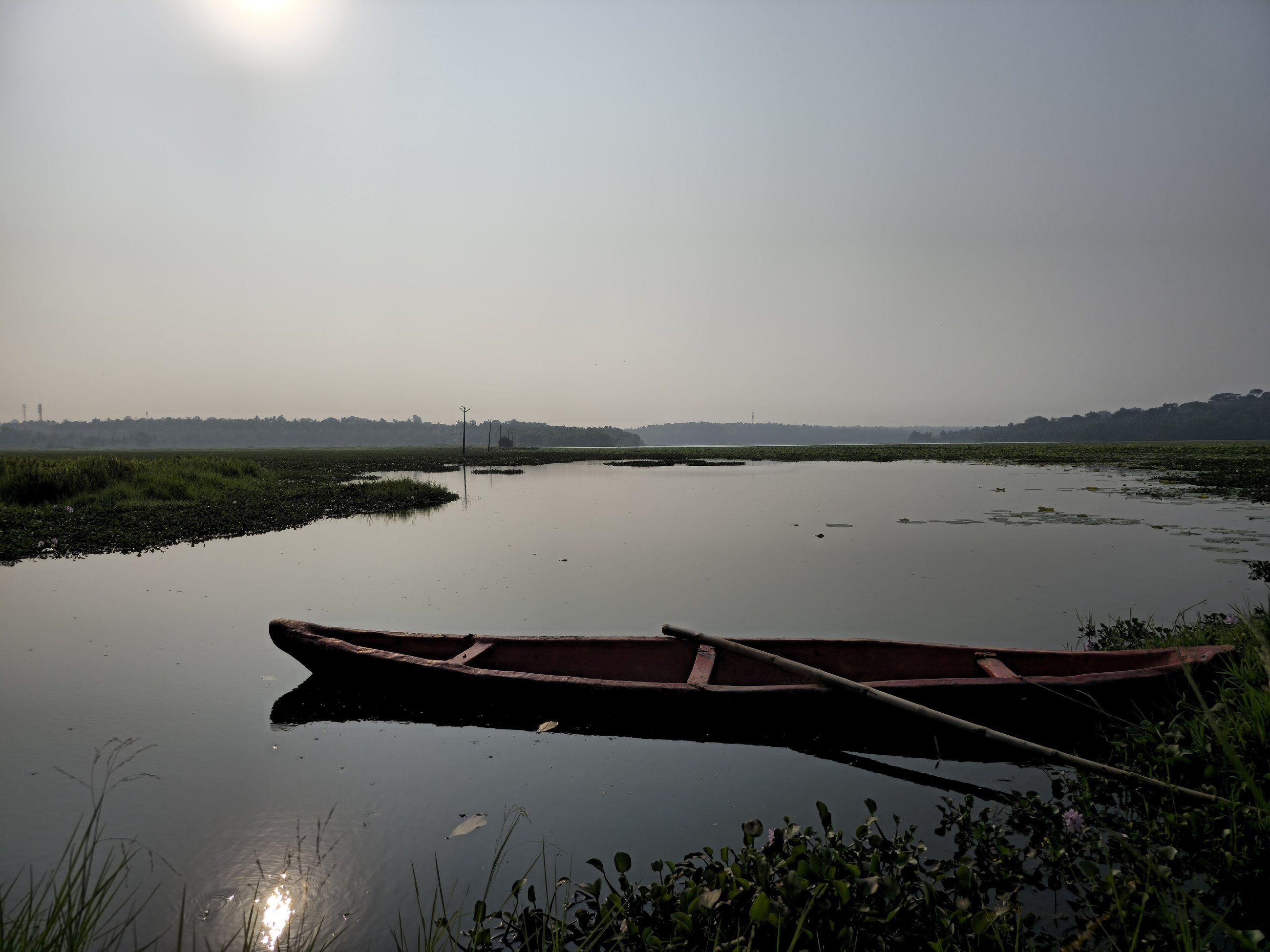 A wooden canoe moored on still water at Vellayani Lake under a hazy sun
